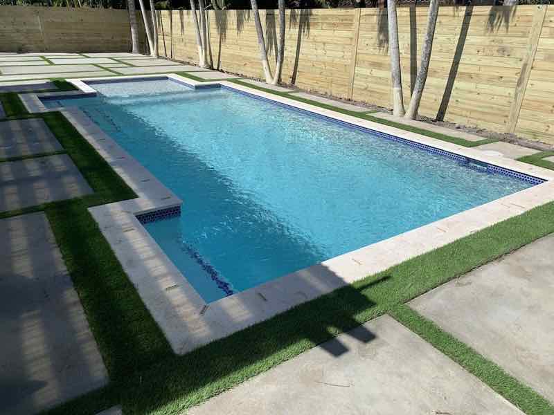Rectangular pool with raised spa set against a turf-and-paver checkerboard deck and wood privacy fence