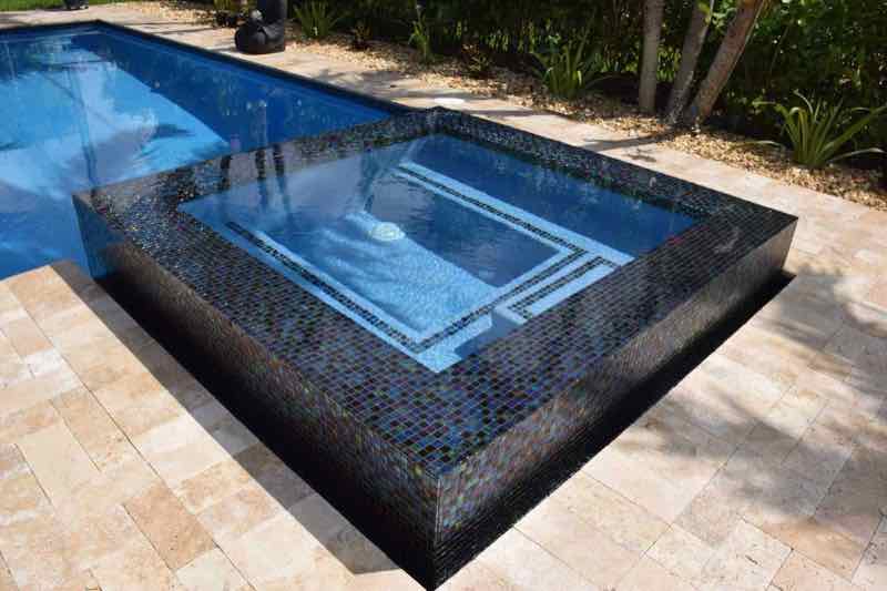 Dark mosaic-tile raised spa with a geometric water feature on a travertine deck