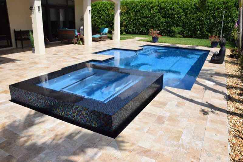 Rectangular pool with dark mosaic raised spa beside a covered patio and travertine deck