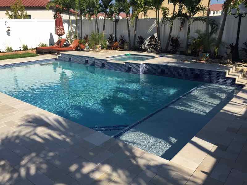 Rectangular pool with raised mosaic-tile spa framed by palm trees beside a tile-roofed home