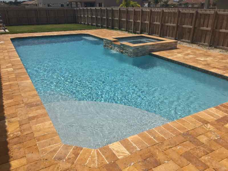 Rectangular pool with raised spa on a brick-paver deck beside a wood privacy fence