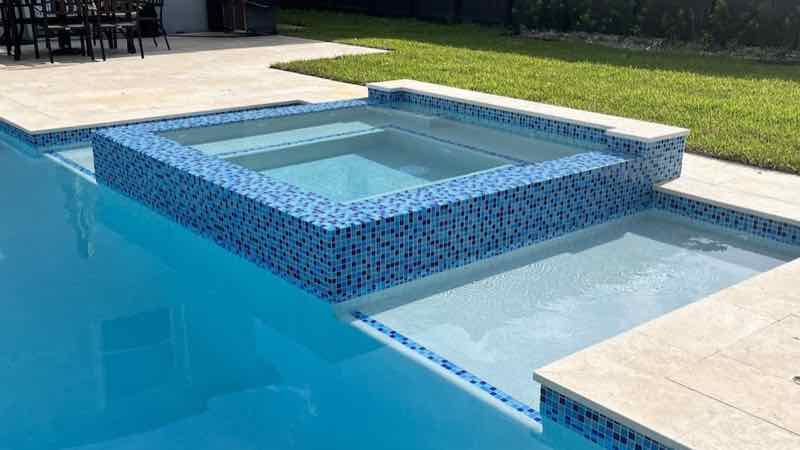 Rectangular pool with raised mosaic-tile spa lit blue on a travertine deck