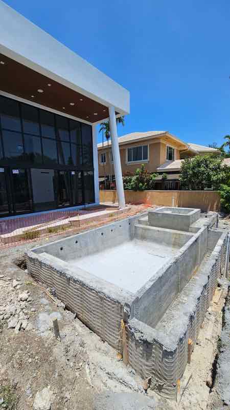Pool shell mid-construction beside a modern home's glass entry