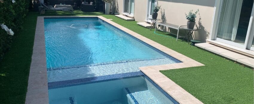 A modern backyard with a rectangular pool featuring a spa section, enhanced by automation systems in Miami-Dade County, FL. The area is surrounded by artificial grass and bordered by a beige patio. On the right, lounge chairs and a small table rest under the "Dream Pools" logo.