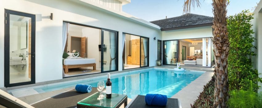 A serene outdoor scene features a private swimming pool with sleek concrete pool decks in Miami-Dade County, FL, surrounded by a modern villa. Two lounge chairs with towels, a table with wine and glasses, and a small green plant bask under the warm natural light.