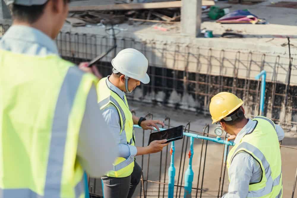 Construction workers wearing safety vests and helmets at a building site are busy with ongoing activities. One examines pipes while another uses a tablet to incorporate automation systems in Miami-Dade County, FL. A third worker is partially visible in the foreground, highlighting the collaborative effort.