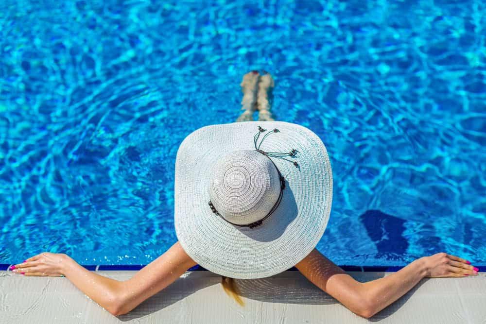 A person in a large white sun hat relaxes in a swimming pool, leaning against the edge with arms outstretched. The blue water shimmers in the sunlight from one of the finest pool constructions Miami-Dade County FL has to offer.