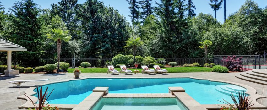 A luxurious backyard in Miami-Dade County, FL, features a large swimming pool with a connected hot tub and elegant concrete pool decks. Surrounded by lush greenery and tall trees, four lounge chairs are placed on the opposite side of the pool. Potted plants decorate the poolside area.