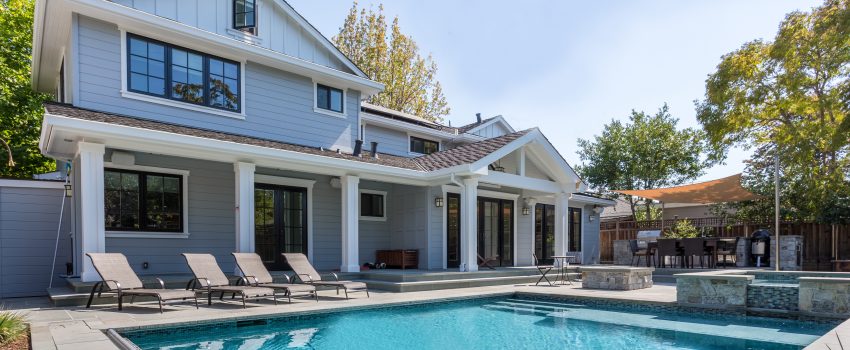 A modern two-story house with a light grey facade and large windows features a spacious backyard. Highlighting its charm, the rectangular swimming pool boasts concrete pool decks Miami-Dade County FL, surrounded by lounge chairs and a shaded sitting area with trees in the background.