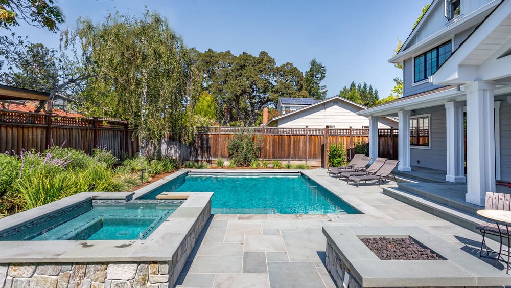 A backyard pool area featuring a swimming pool and hot tub with concrete pool decks in Miami-Dade County, FL. Surrounded by stone tiles, there are lounge chairs nearby and a fire pit in the foreground. The space is enclosed by a wooden fence, bordered by trees and a house on the right.