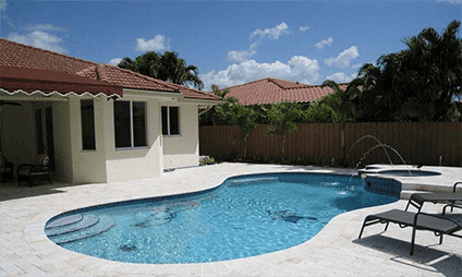 A backyard featuring a kidney-shaped swimming pool with clear blue water under a sunny sky. Two lounge chairs sit on the white-tiled deck near a small hot tub with fountains. Palm trees and a wooden fence line the area, adjacent to a beige house.