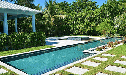 A serene outdoor area with a rectangular swimming pool surrounded by lush greenery and tropical plants. There is a shaded area with a roof on the left and a sunlit patio with stepping stones on the right.