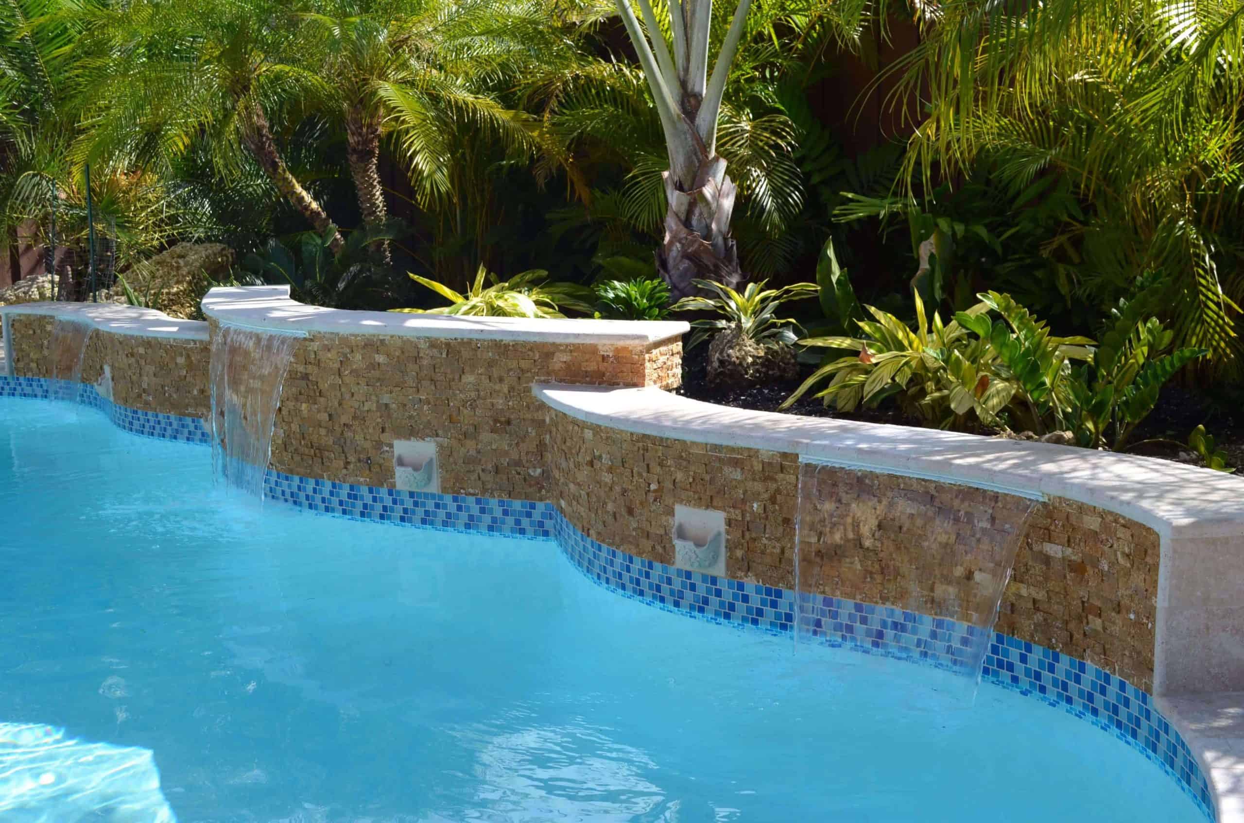 Blue swimming pool with small waterfalls flowing from a stone-tiled wall, crafted by expert pool builders Miami-Dade County, FL, and surrounded by lush tropical plants and palm trees in the background.