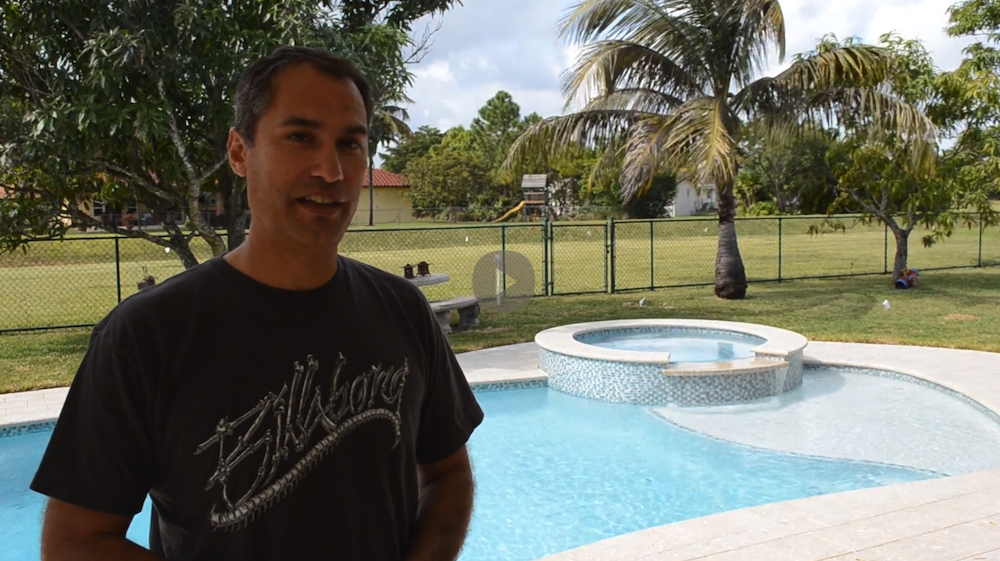 A man stands outdoors near a swimming pool and hot tub, crafted by expert pool builders Miami-Dade County, with a lush lawn, palm tree, and playground in the background on a sunny FL day.