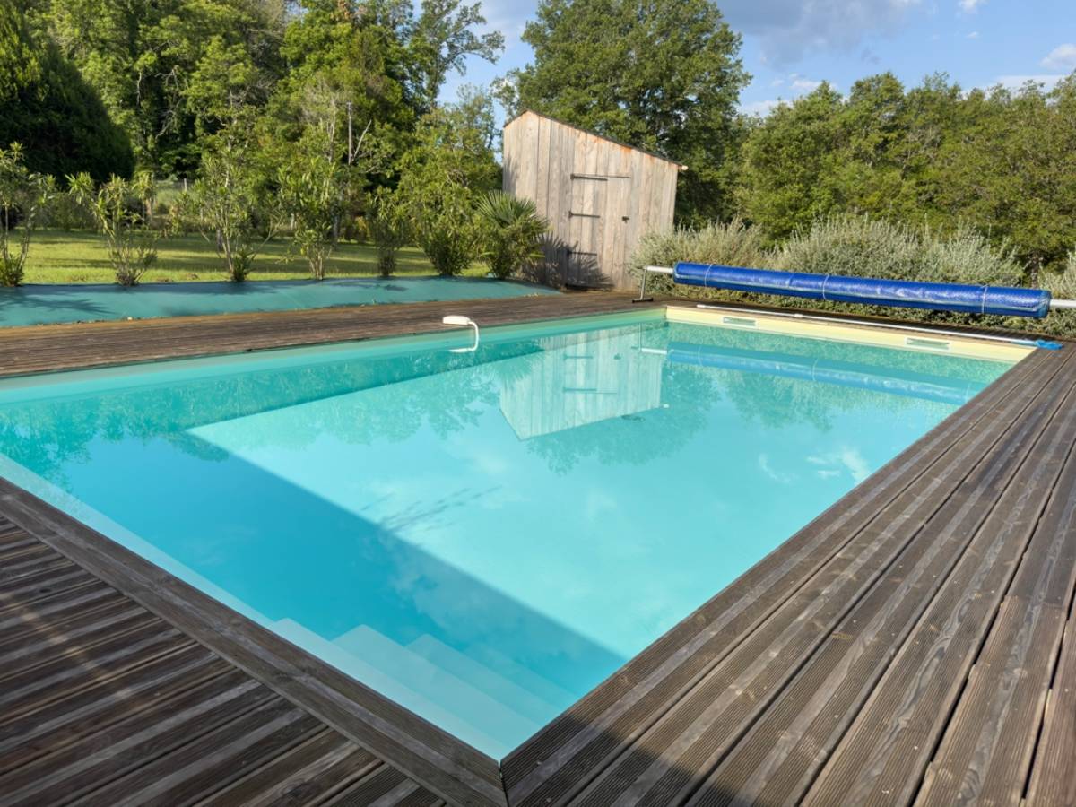 A rectangular outdoor swimming pool with clear blue water, crafted by top pool builders in Miami-Dade County, is surrounded by a wooden deck. Trees and greenery frame the scene, along with a small wooden shed and a blue pool cover roller.