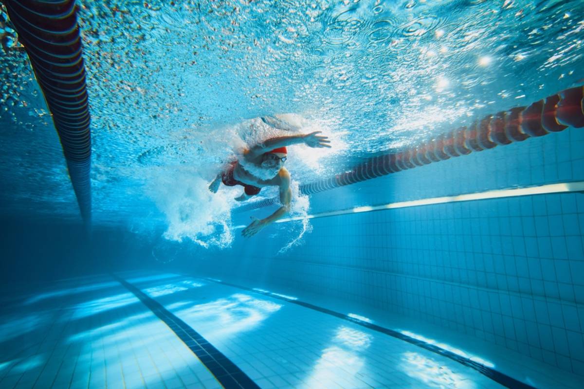 Underwater view of a swimmer in a clear blue pool, built by expert pool builders Miami-Dade County, wearing goggles and a swim cap as they create splashes swimming freestyle near a red lane divider. Sunlight filters through the water.