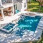 Aerial view of a modern backyard in Miami-Dade County, FL, showcasing expert pool construction with a rectangular swimming pool, attached hot tub, outdoor kitchen, dining area, lounge chairs on a stone patio beside a grassy lawn and palm tree.