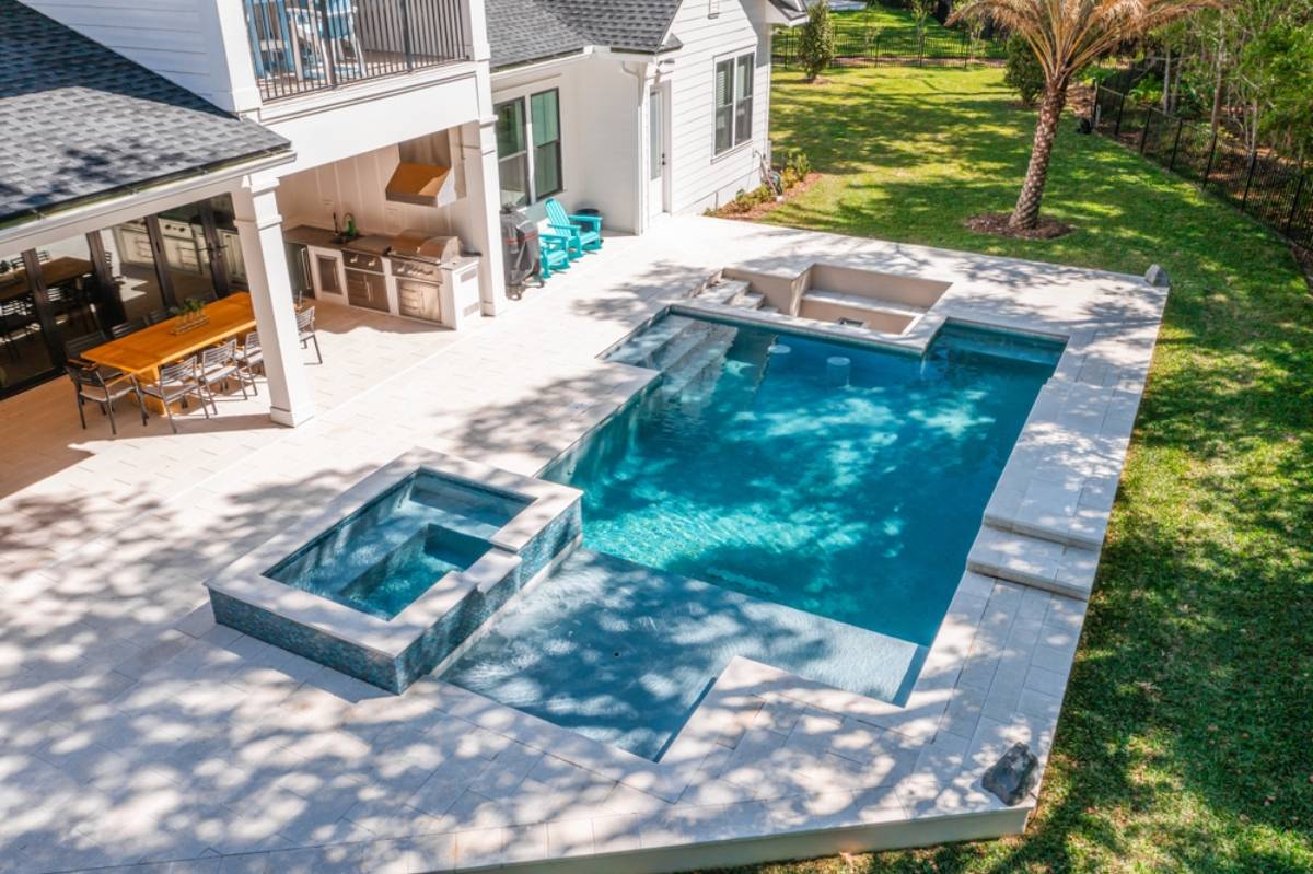 Aerial view of a modern backyard in Miami-Dade County, FL, showcasing expert pool construction with a rectangular swimming pool, attached hot tub, outdoor kitchen, dining area, lounge chairs on a stone patio beside a grassy lawn and palm tree.