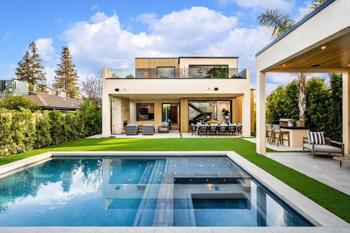 Modern two-story house in FL with large windows, patio seating, outdoor dining area, and a swimming pool with a spa—featuring expert pool construction Miami-Dade County, FL—surrounded by green lawn and privacy hedges under a blue sky.